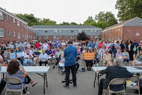 Several hundred residents seated and standing in the courtyard at Bell Park Manor Terrace, listening to a speaker, many holding Co-ops & Condos United signs